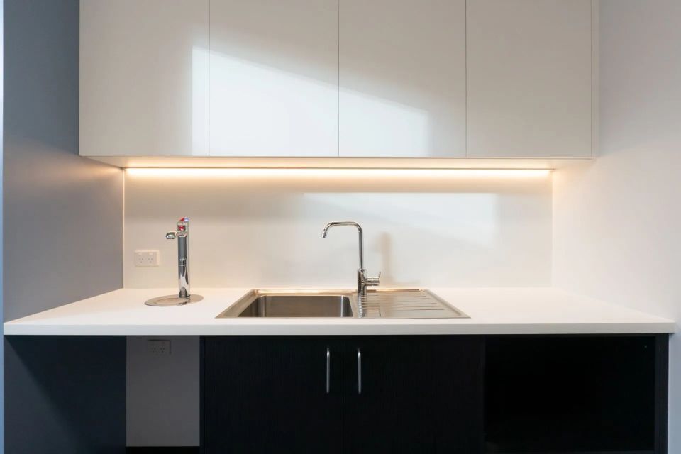 Kitchen sink area with stainless steel faucet and white countertops, underlit by a strip light.