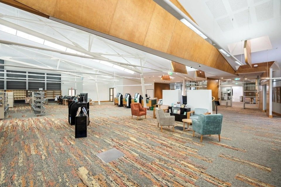 Spacious library interior with bookshelves, seating areas, and a patterned carpet. Natural light streams in from large windows.