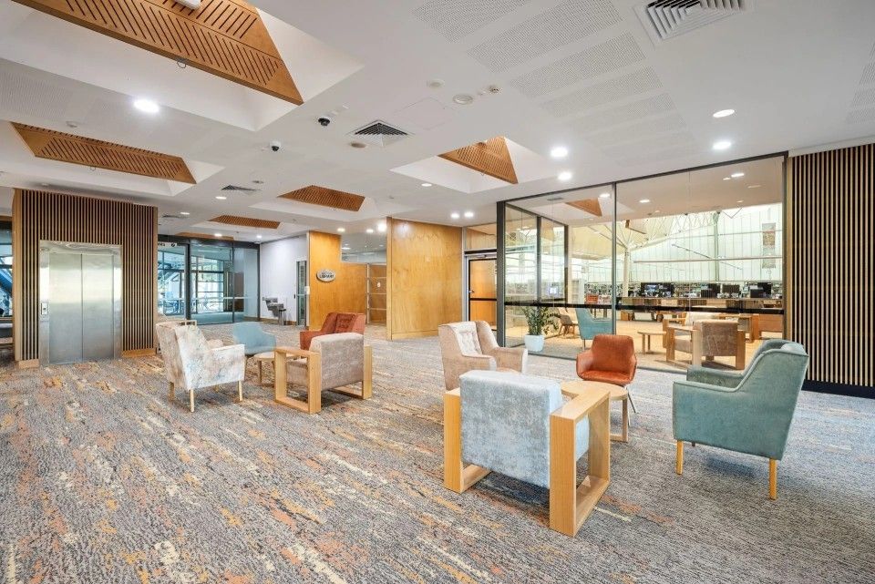 Lounge area with armchairs and small tables on patterned carpet. Wooden accents, windows, and bright lighting.