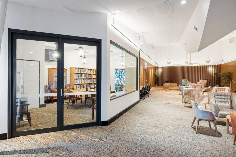 Interior with glass doors, leading to library; seating area with patterned chairs, neutral carpet, and lighting.