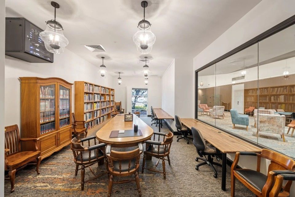 Library interior with bookshelves, tables, and chairs. Large window offers view of lounge.