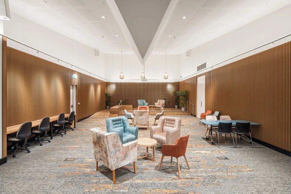 A large, modern room with seating, tables, and a long desk along the wall. Wooden panels and patterned carpet.