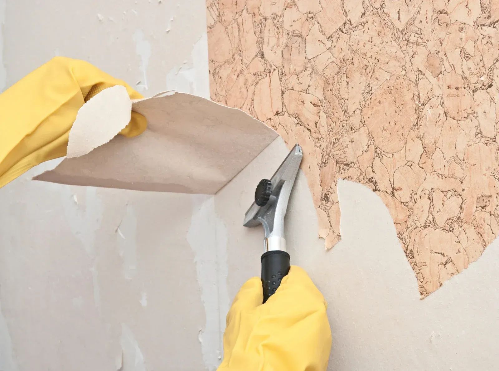 Hands in yellow gloves scrape peeling wallpaper with a putty knife on a wall