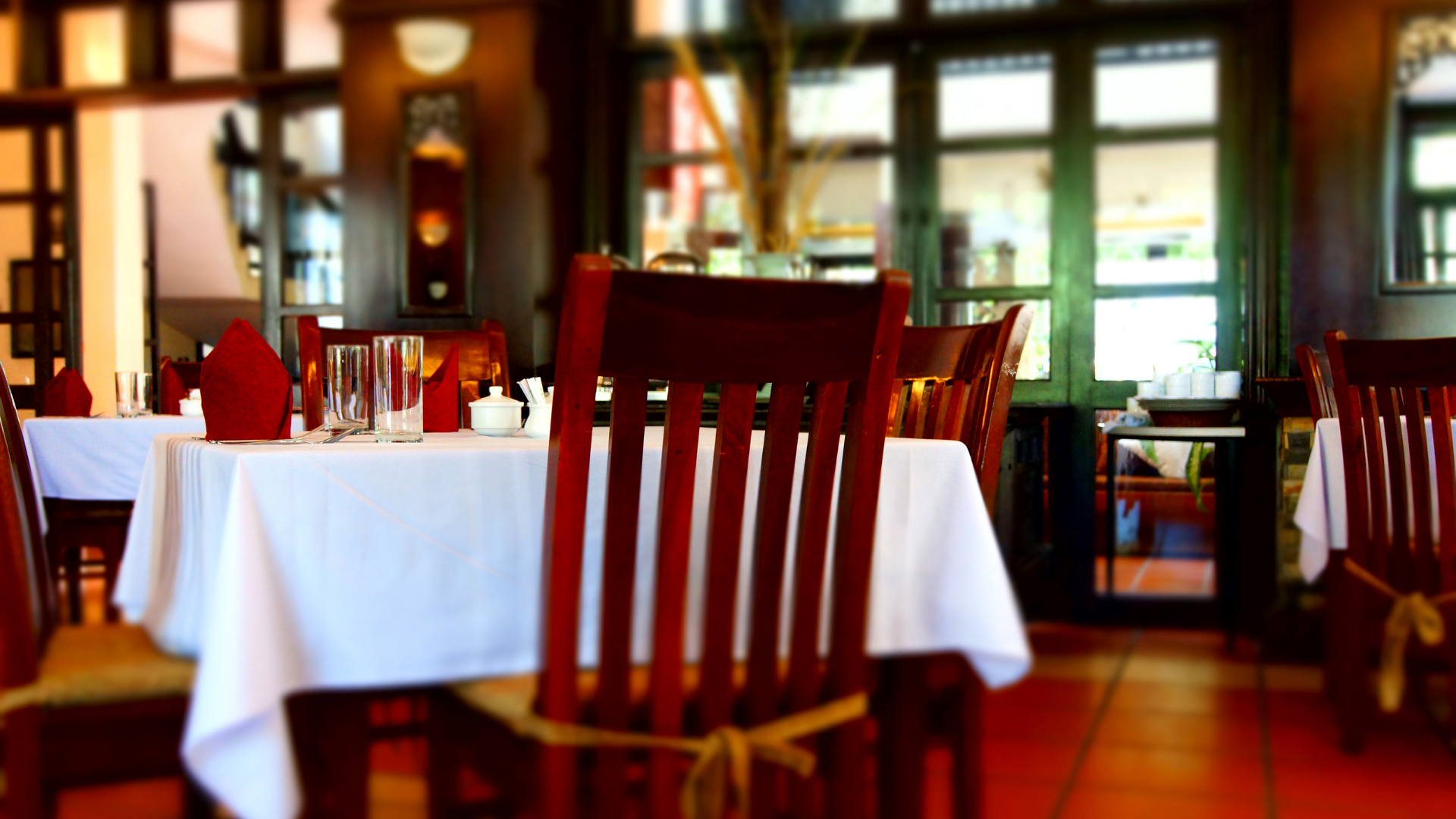 Empty restaurant dining room with white tablecloths and dark wooden chairs, softly lit by warm windows