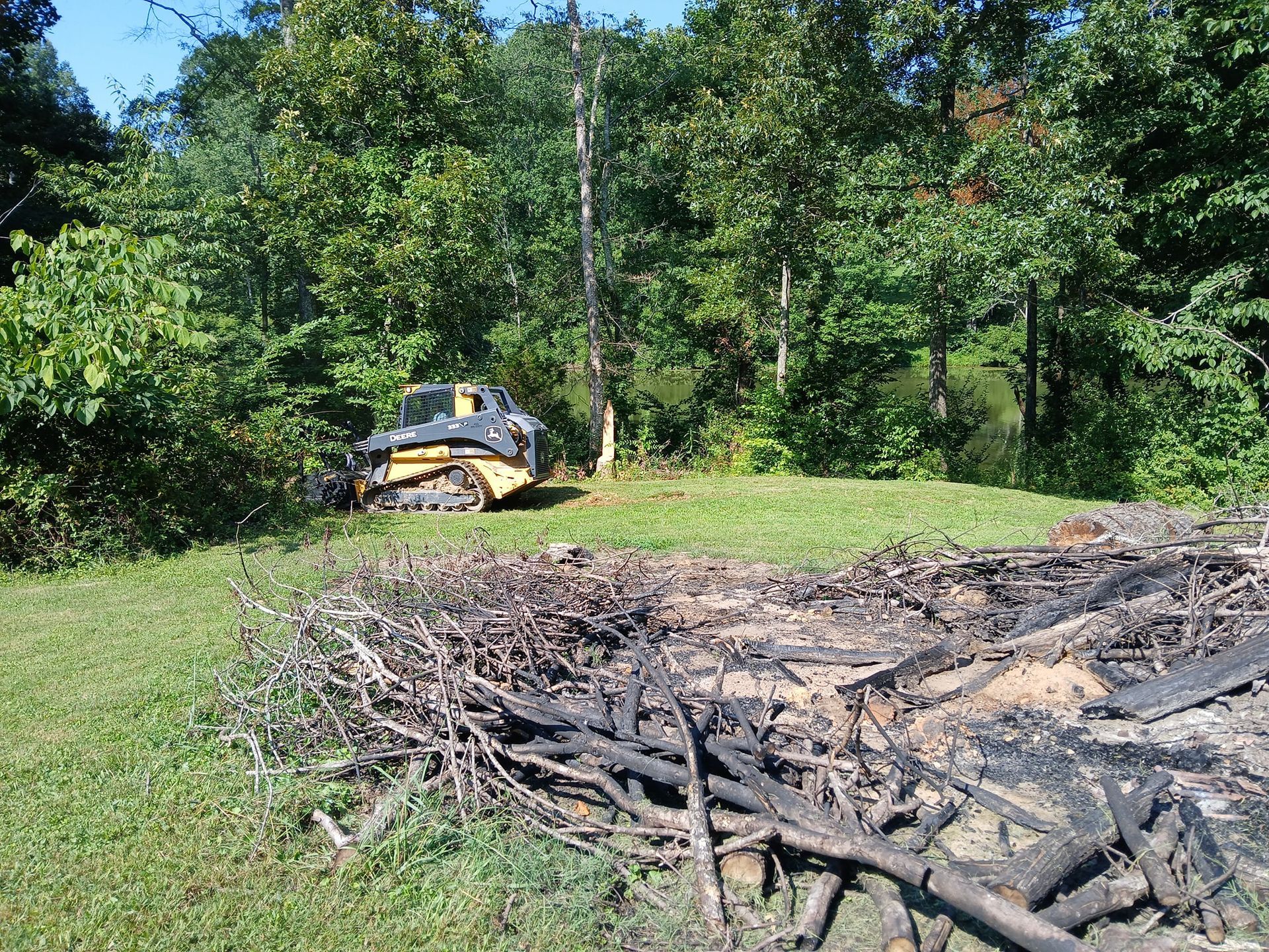 A bulldozer clearing a grassy area near a pile of burnt debris, with trees in the background.