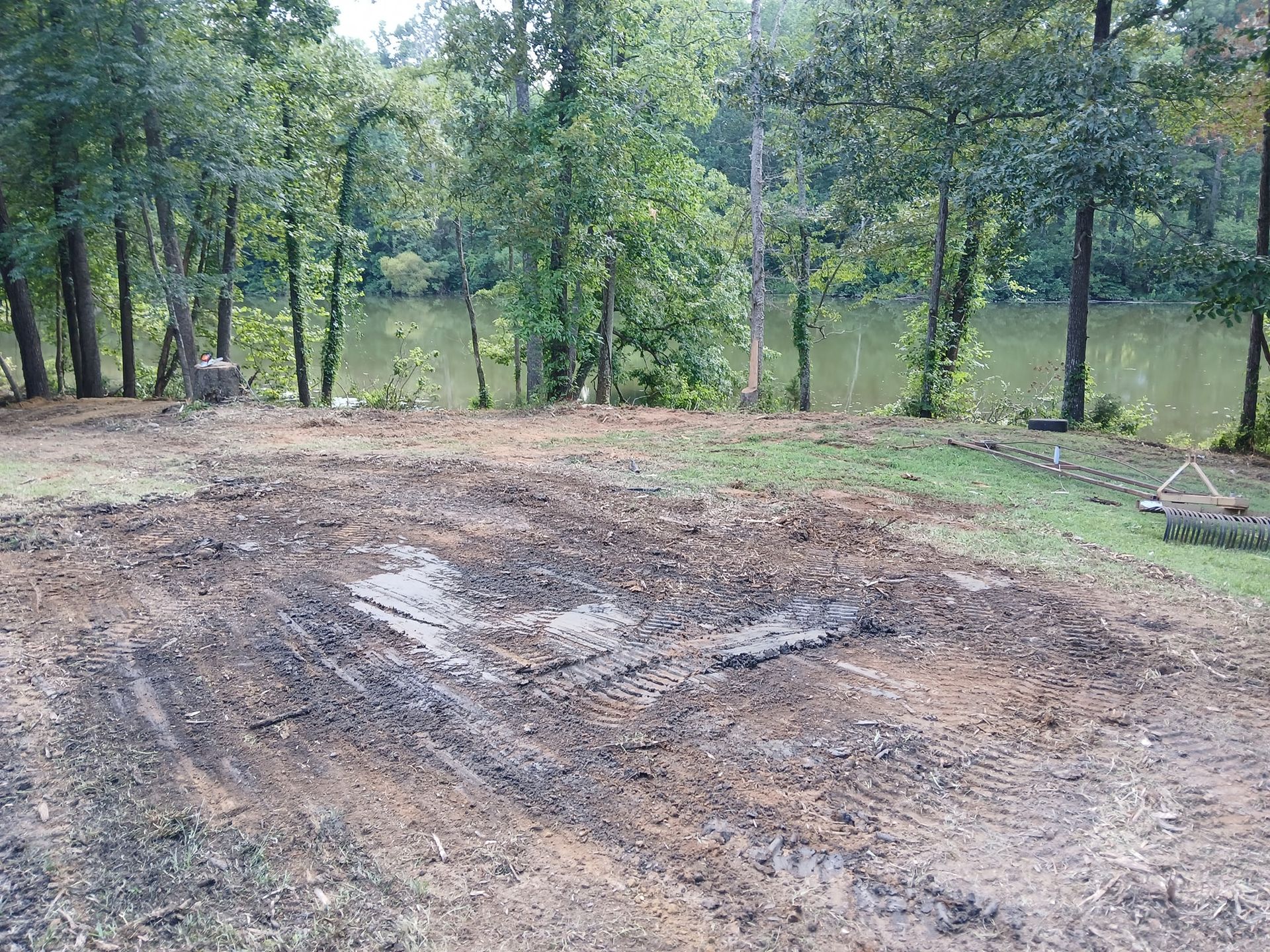 Dirt clearing with water puddle, surrounded by grass, trees, and a lake in the background.