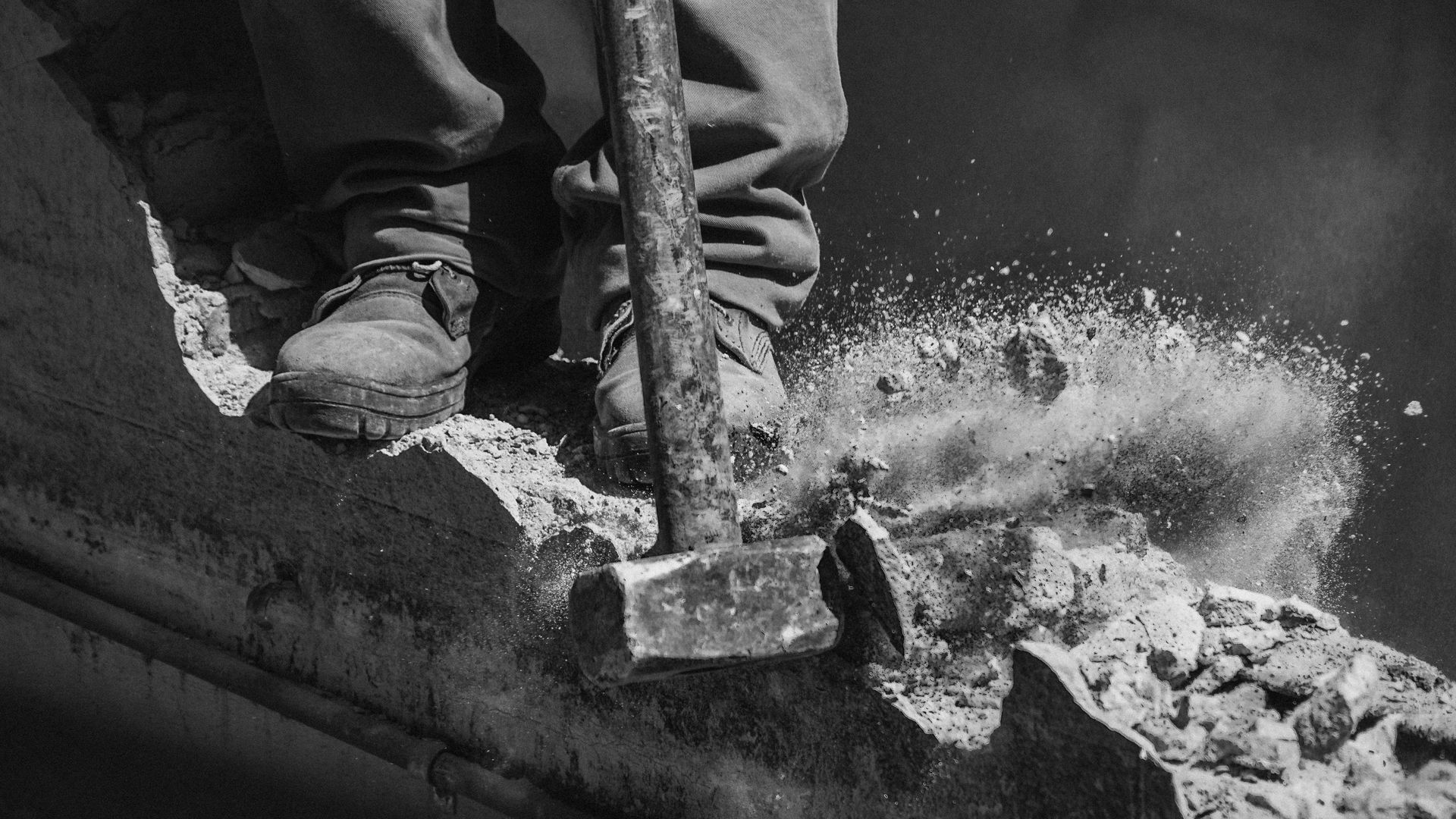 Worker's legs and feet atop concrete, smashing with a hammer, causing dust to fly.