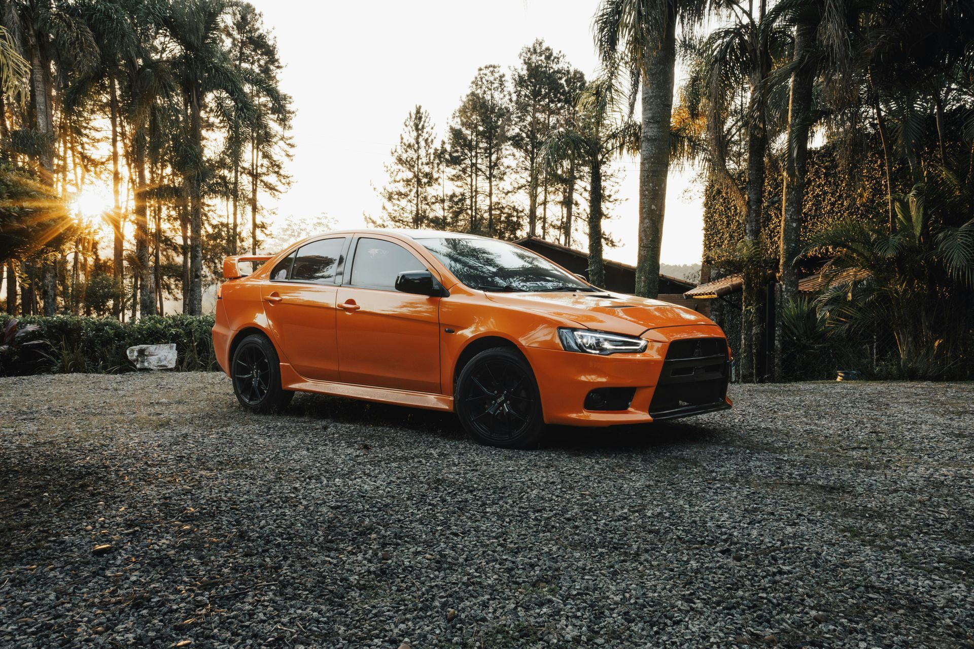 Orange sports car parked on gravel, trees in background, sunset.