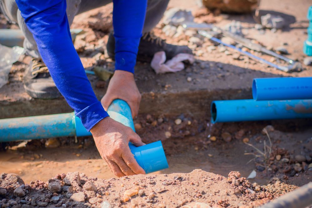 A plumber connecting blue PVC pipes in a dirt trench outdoors.