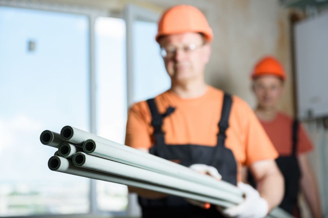 Construction worker holding pipes, orange hat, overalls. Another worker in the background.