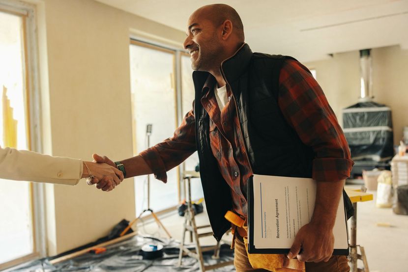Man in work clothes shaking hands, smiling, holding contract. Interior with tools and construction materials.
