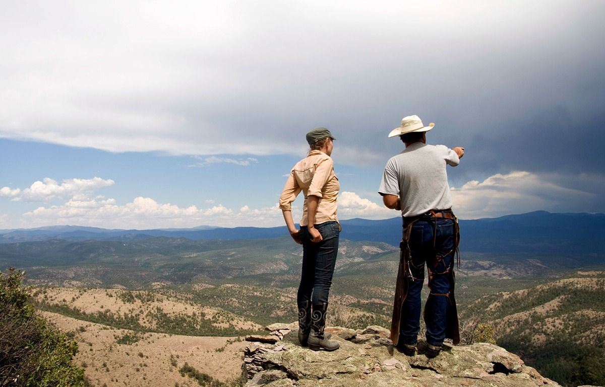 Two men on a cliff overlook a mountain range; one points, and the sky is partly cloudy.