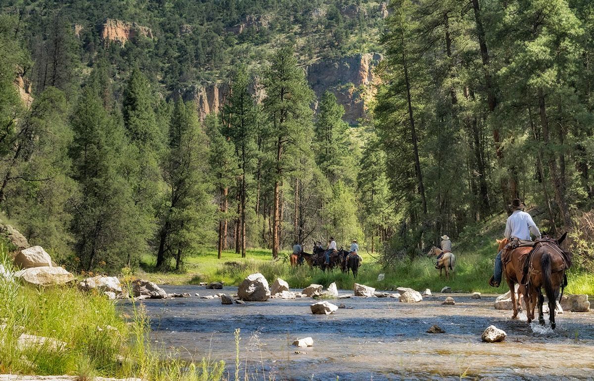 People on horseback crossing a shallow, rocky river through a forest.