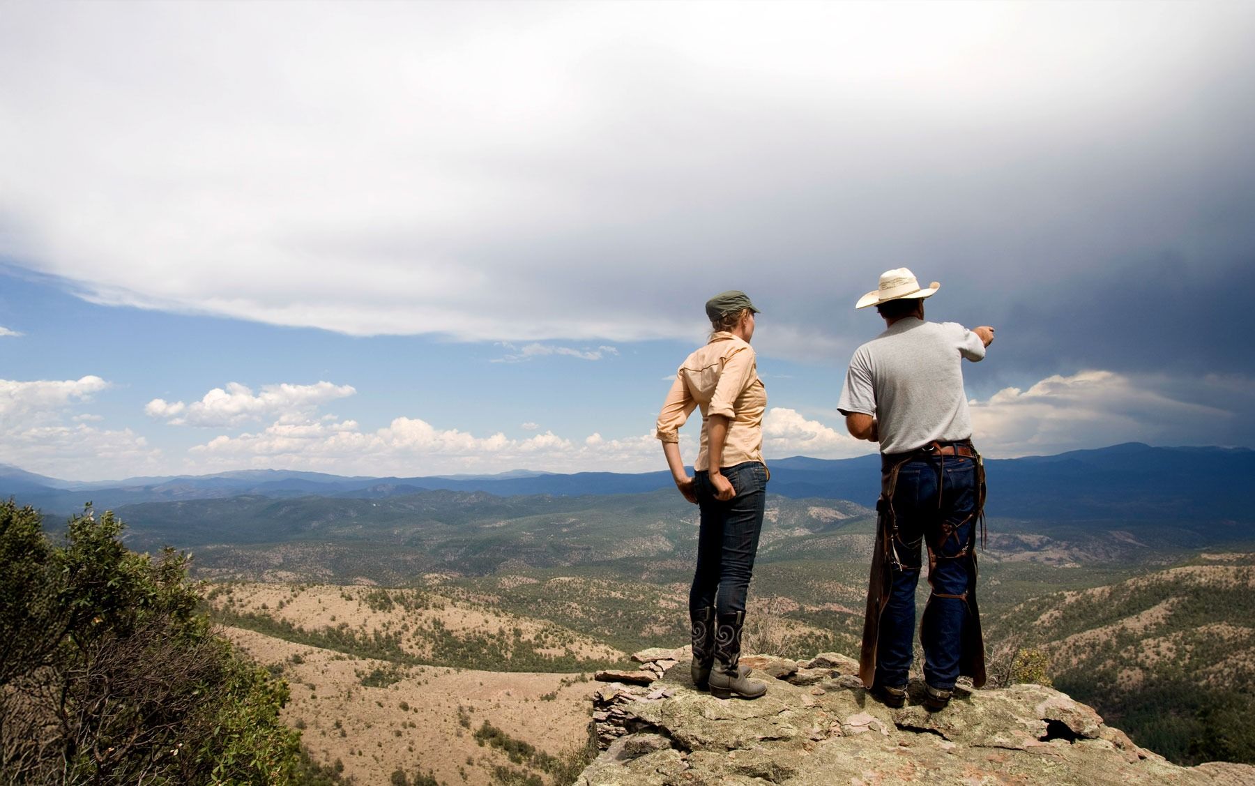Two people on a rocky cliff, looking at a valley. One points toward distant mountains. Cloudy sky.