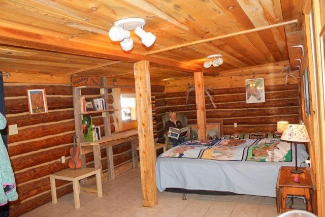 Cozy log cabin bedroom with wooden beams, bed, desk, person reading, and warm lighting.