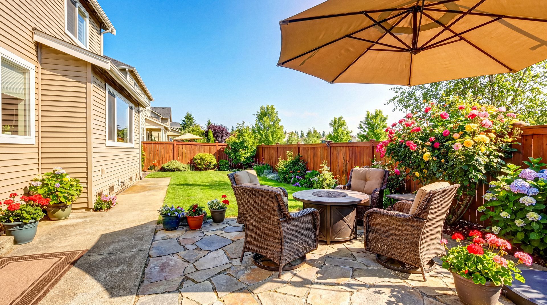 Outdoor patio with wicker chairs, umbrella, colorful flowers, and a landscaped backyard garden.