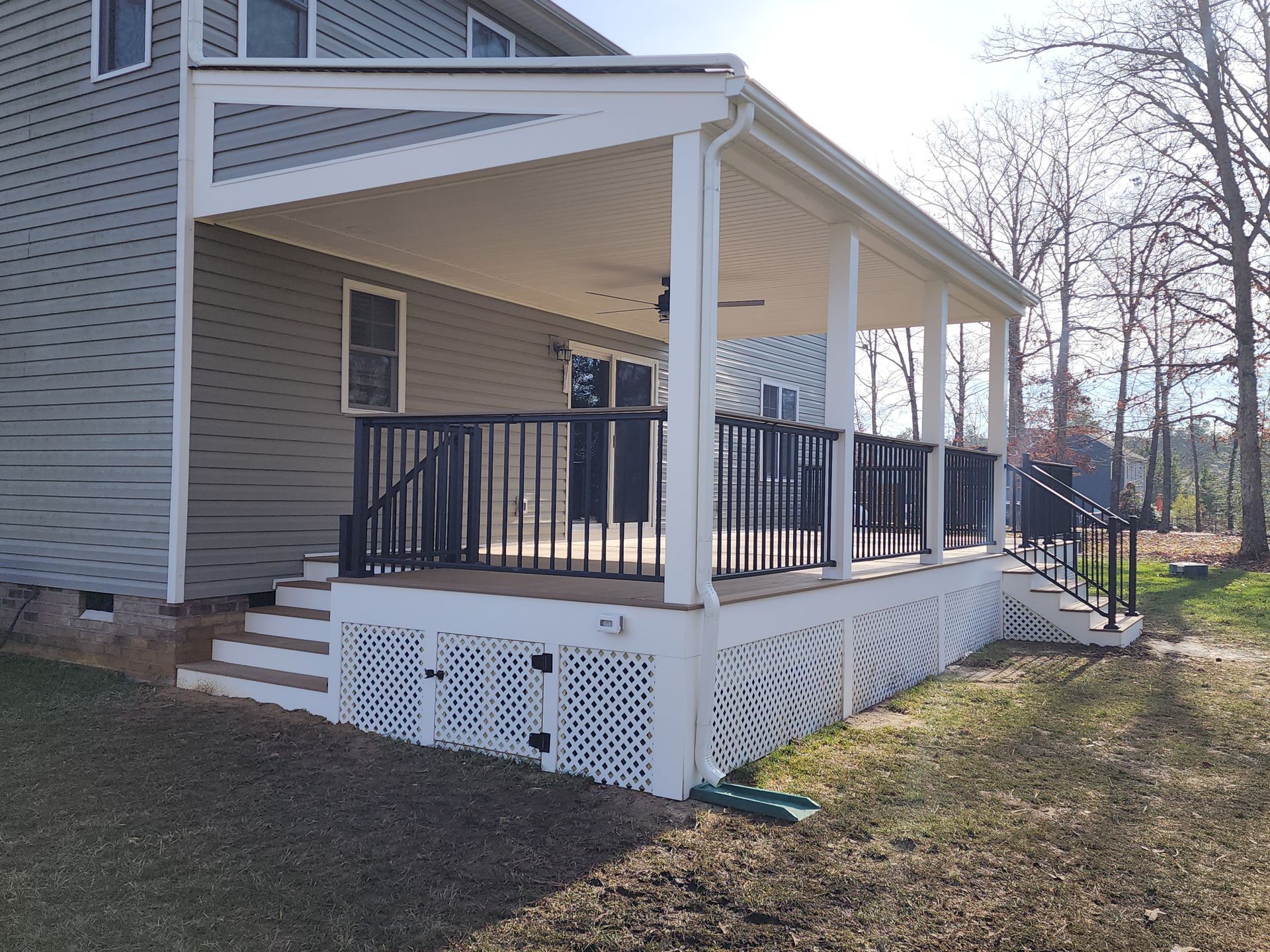 Covered deck with white lattice skirt, black railing, and steps leading to a house with gray siding.