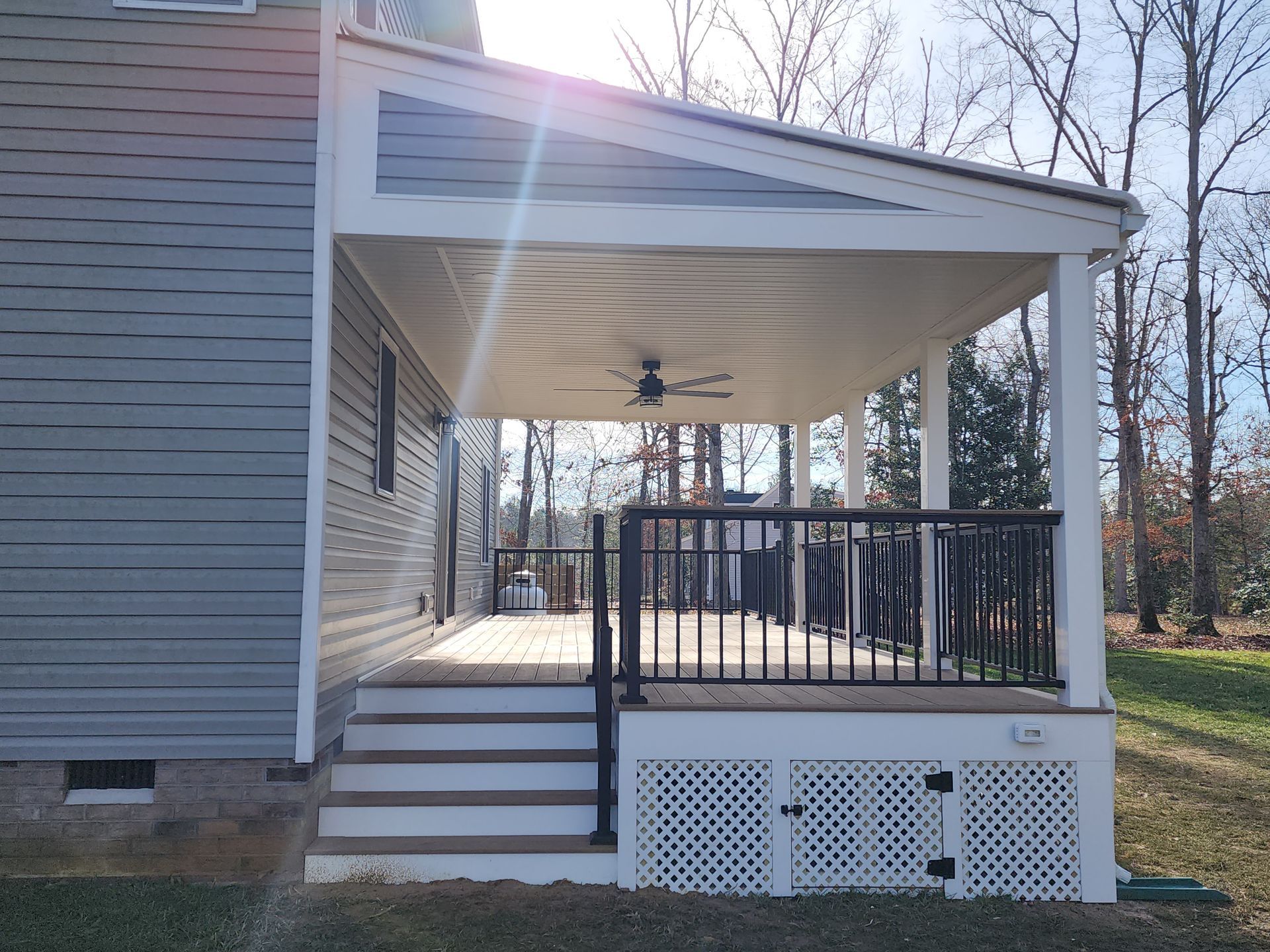Covered deck with stairs, railing, and a ceiling fan, attached to a gray house, in a grassy yard.