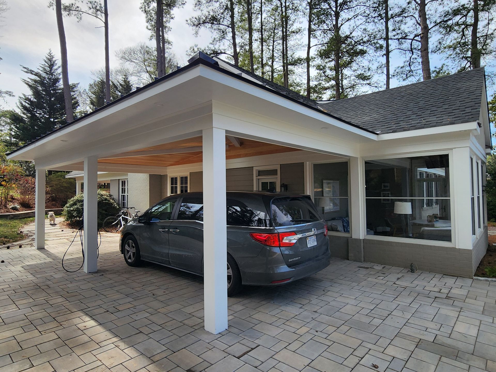 Gray minivan parked under a white carport connected to a house with a gray roof and glass windows on a brick driveway.