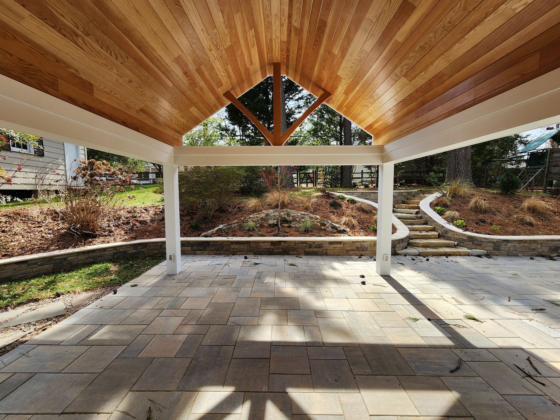 Patio with stone floor, wood ceiling, white columns, overlooking a landscaped yard.