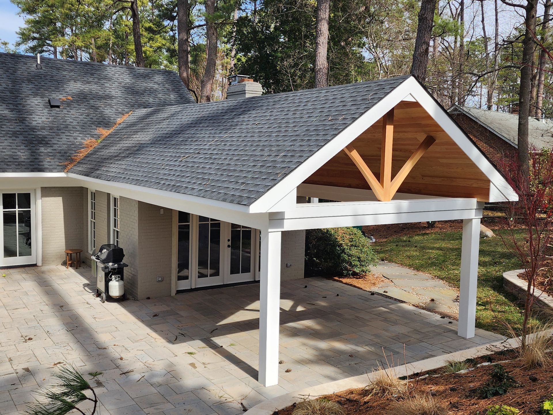 White-columned covered patio attached to a house with a gray roof.  A wooden beam is visible.