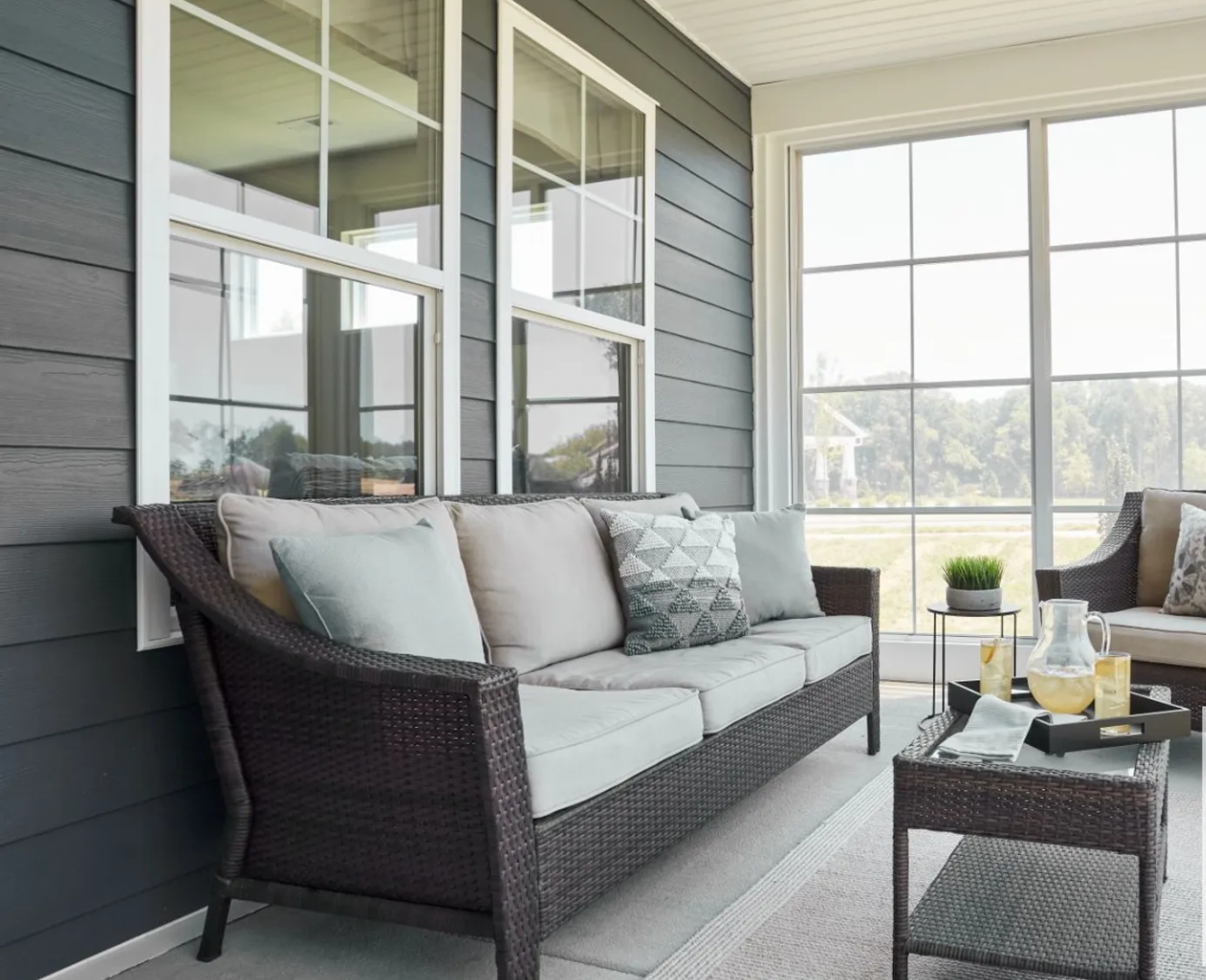 Wicker sofa with blue and cream pillows on a screened porch with gray siding and large windows.