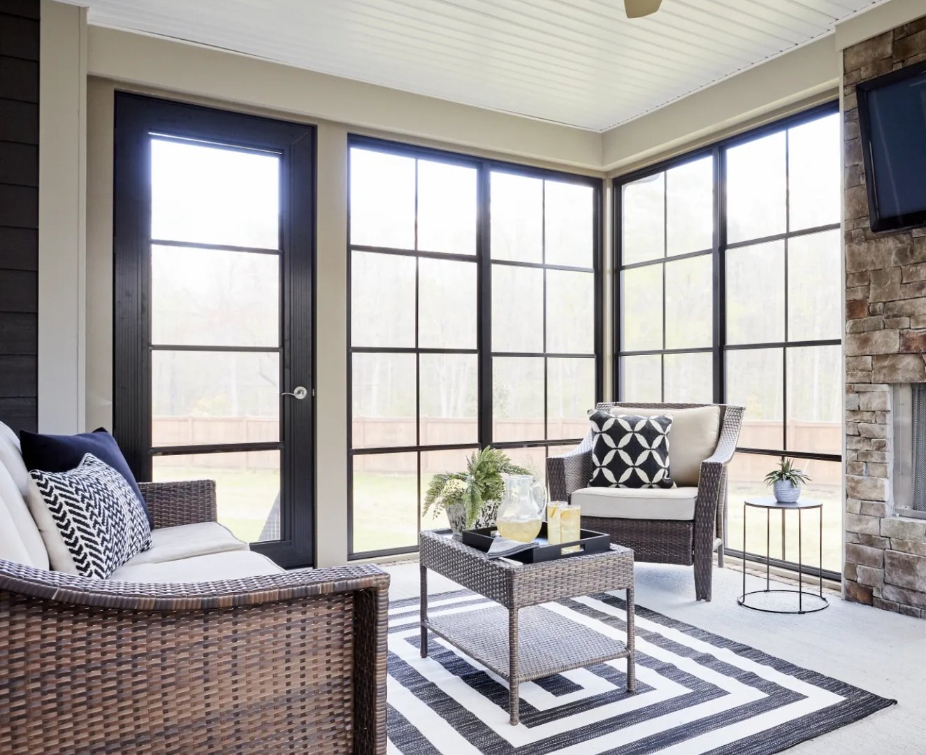 Sunroom with wicker furniture, a black and white rug, and large windows.