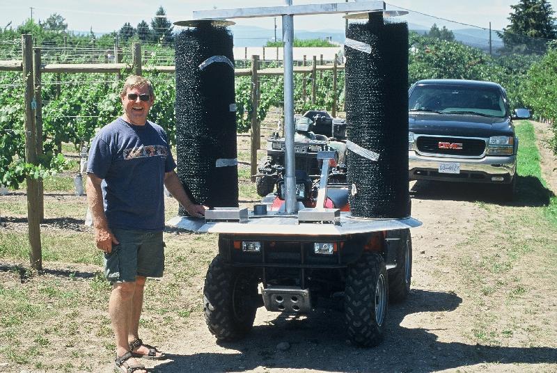 Man standing next to ATV with vertical brushes used for vineyard maintenance. Sunny day.