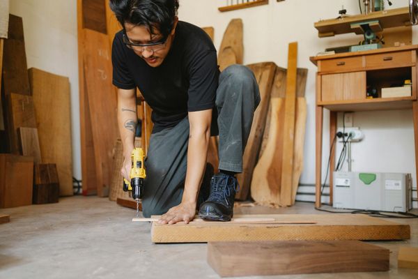 Man in a workshop using a drill on wood. He kneels on the floor, surrounded by lumber.