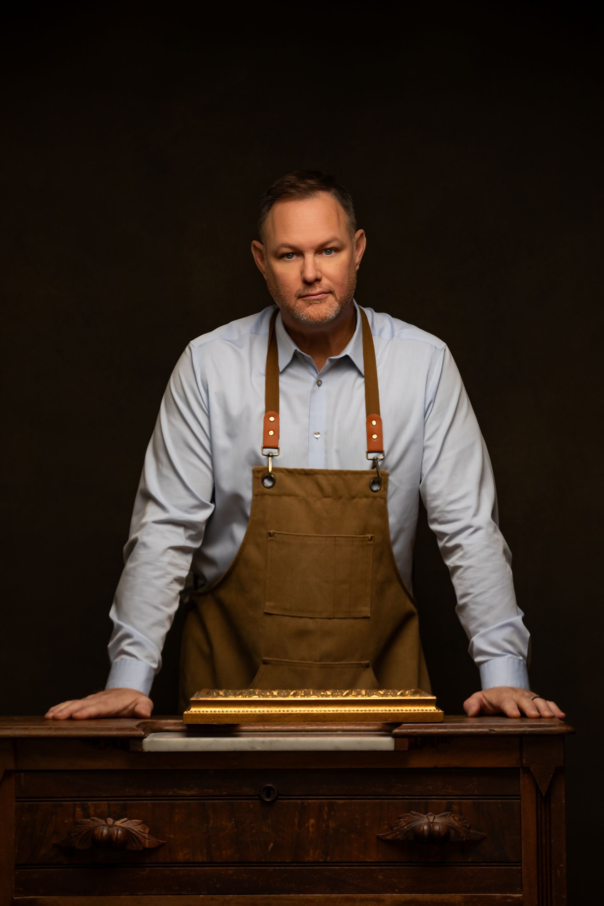 Man in a white shirt and brown apron standing behind a wooden workbench against a dark background.