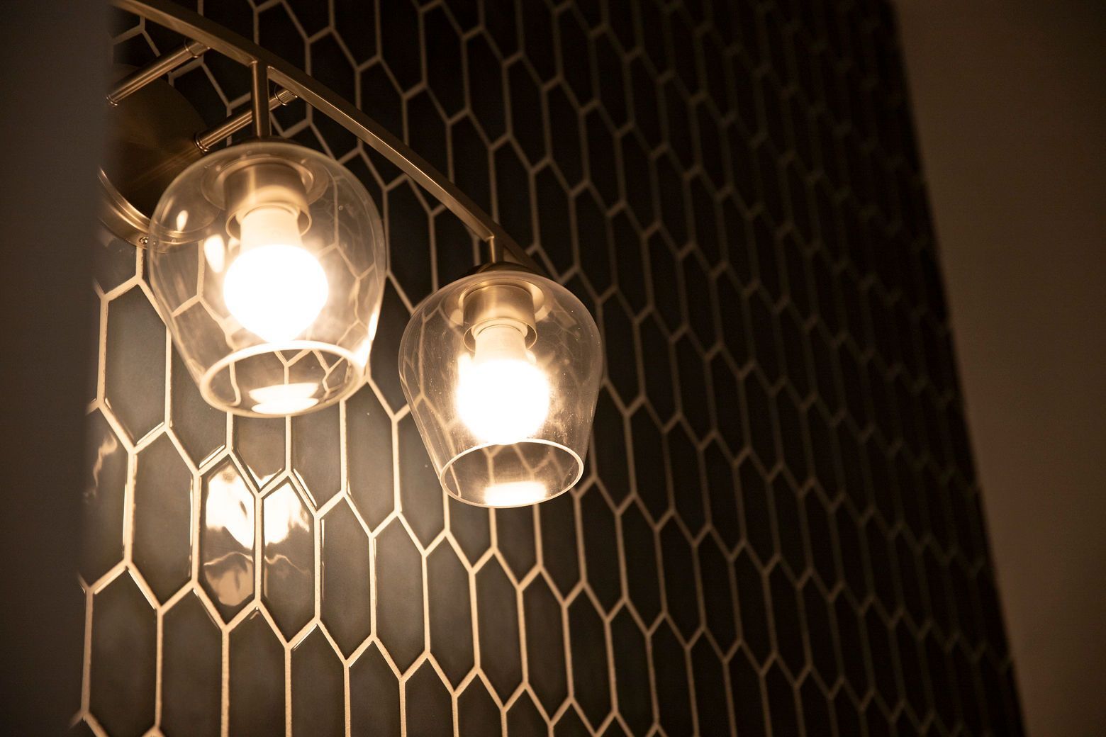 A modern light fixture with a gold-toned finish against the dark hexagonal tile accent wall.
