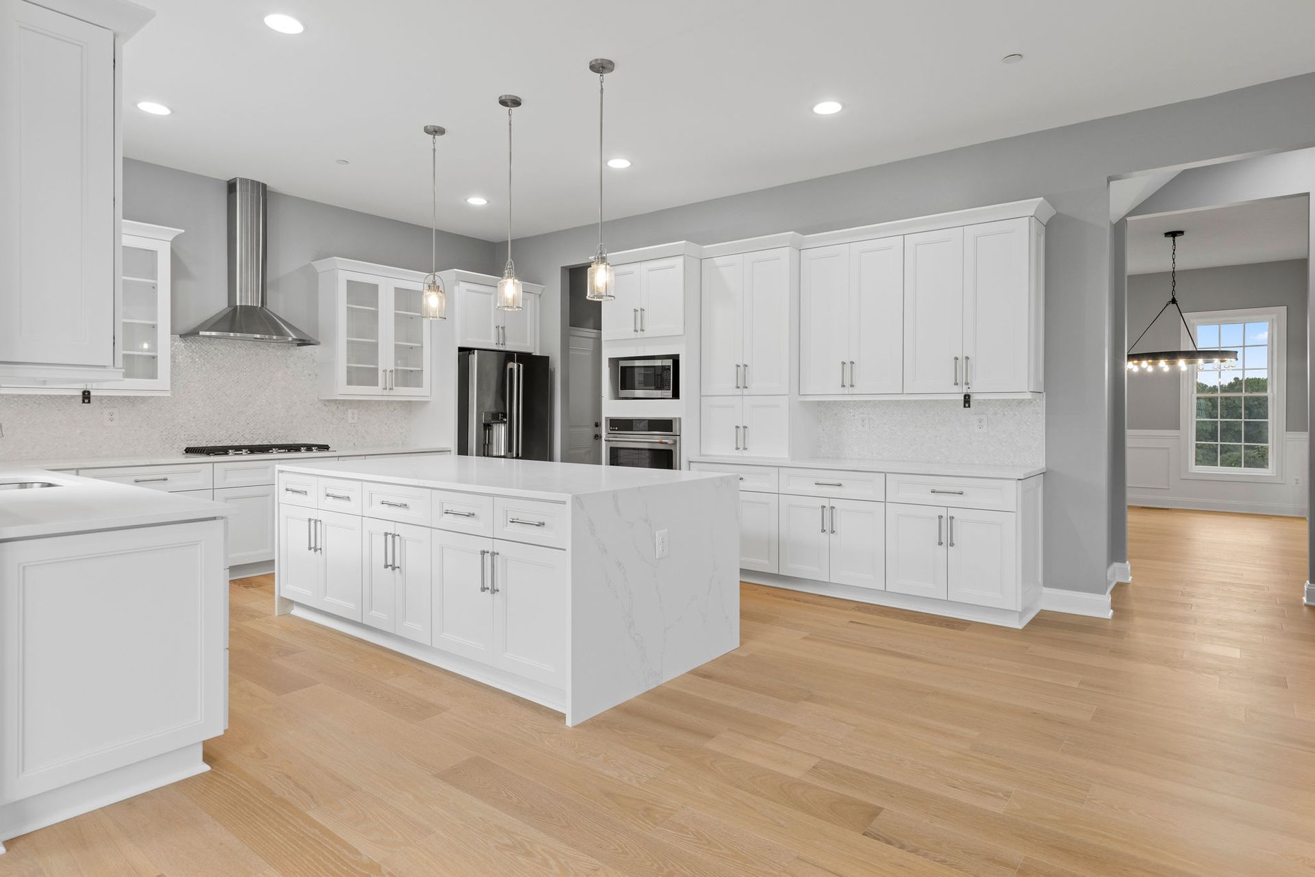 A brightly lit, modern kitchen featuring white shaker cabinets, a large central island with seating, and gleaming hardwood floors.