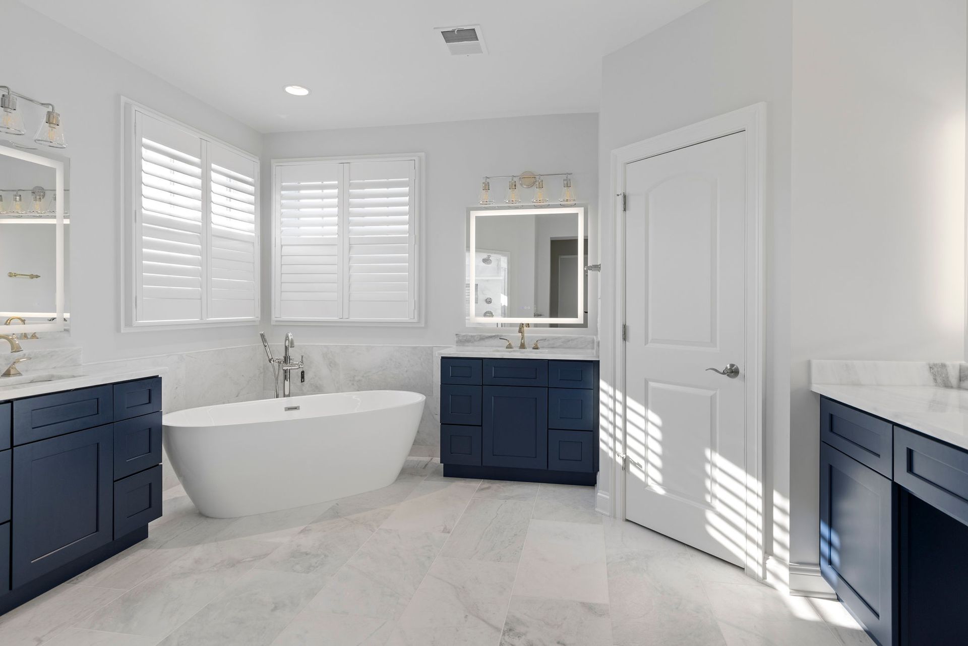 A bright and modern master bathroom with a walk-in shower, elegant blue vanities, and a freestanding tub.