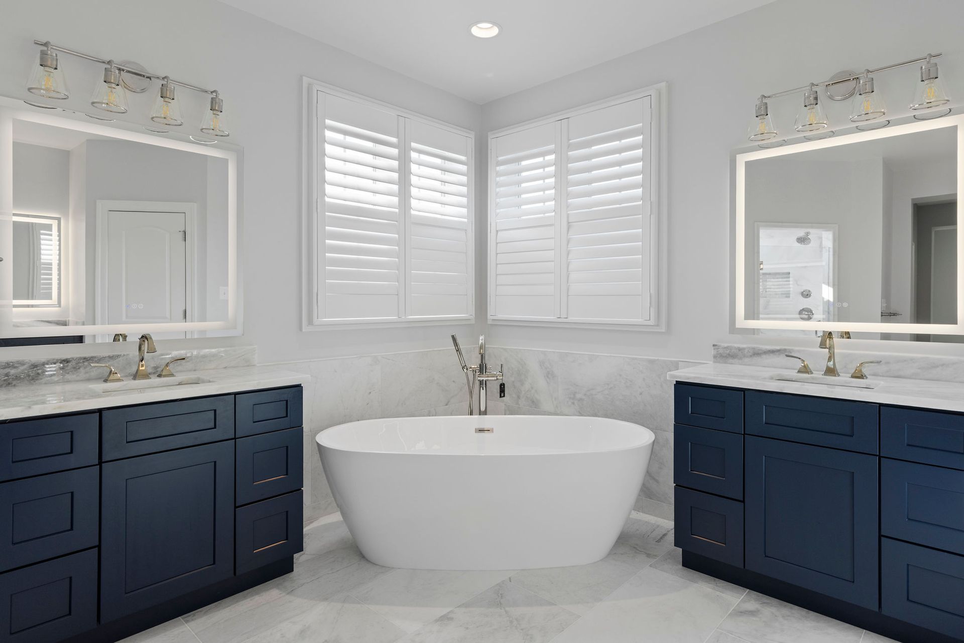 Modern bathroom renovation in Baltimore, MD with navy blue double vanities, marble-style tile walls, and a central white soaking tub.