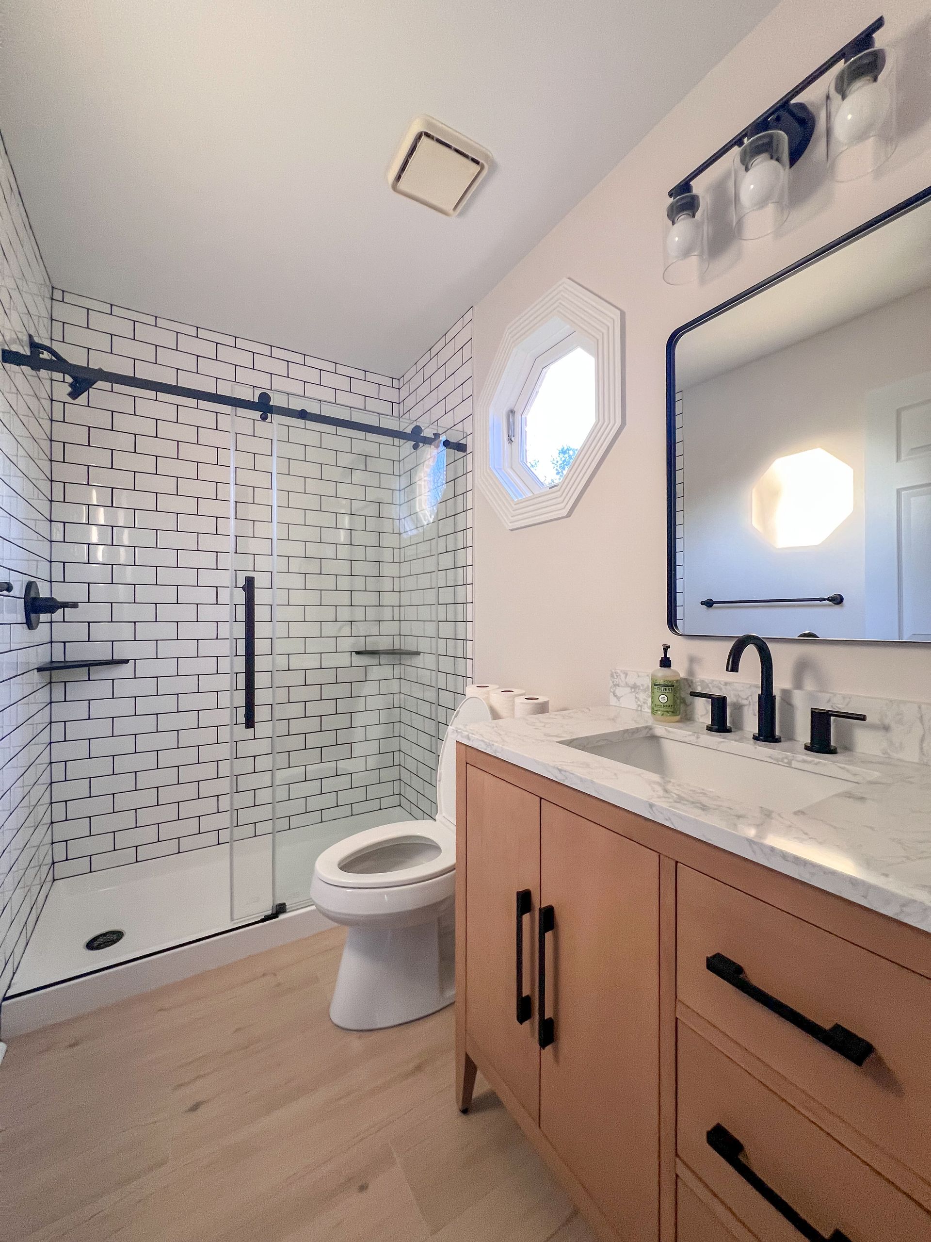 Renovated bathroom with walk-in shower, white subway tile, light wood vanity, black accents, and glass shower enclosure.