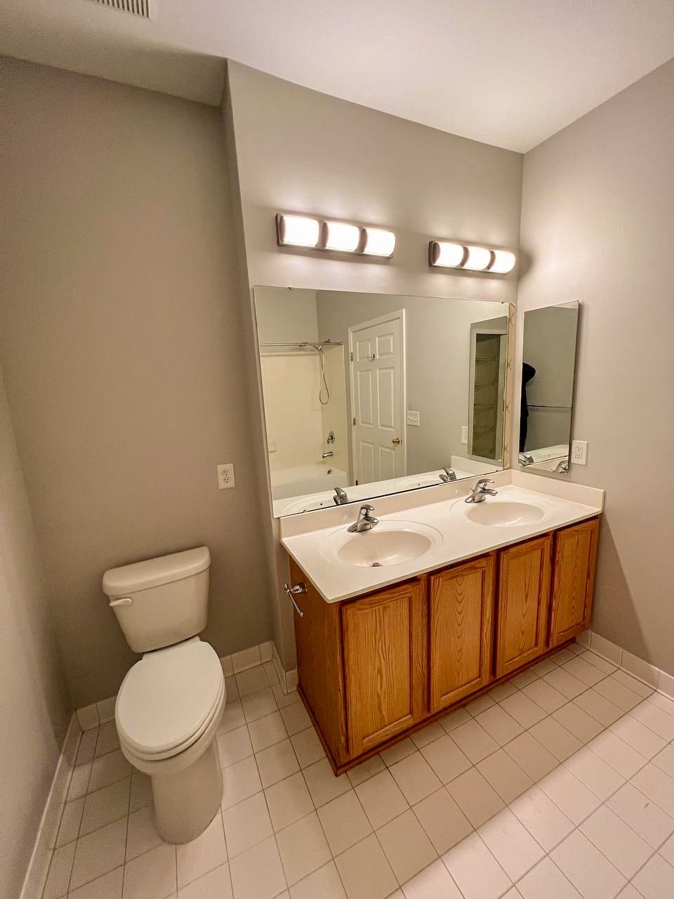 Dated oak vanity with double sinks, white countertop, and large mirror in a bathroom before renovation.