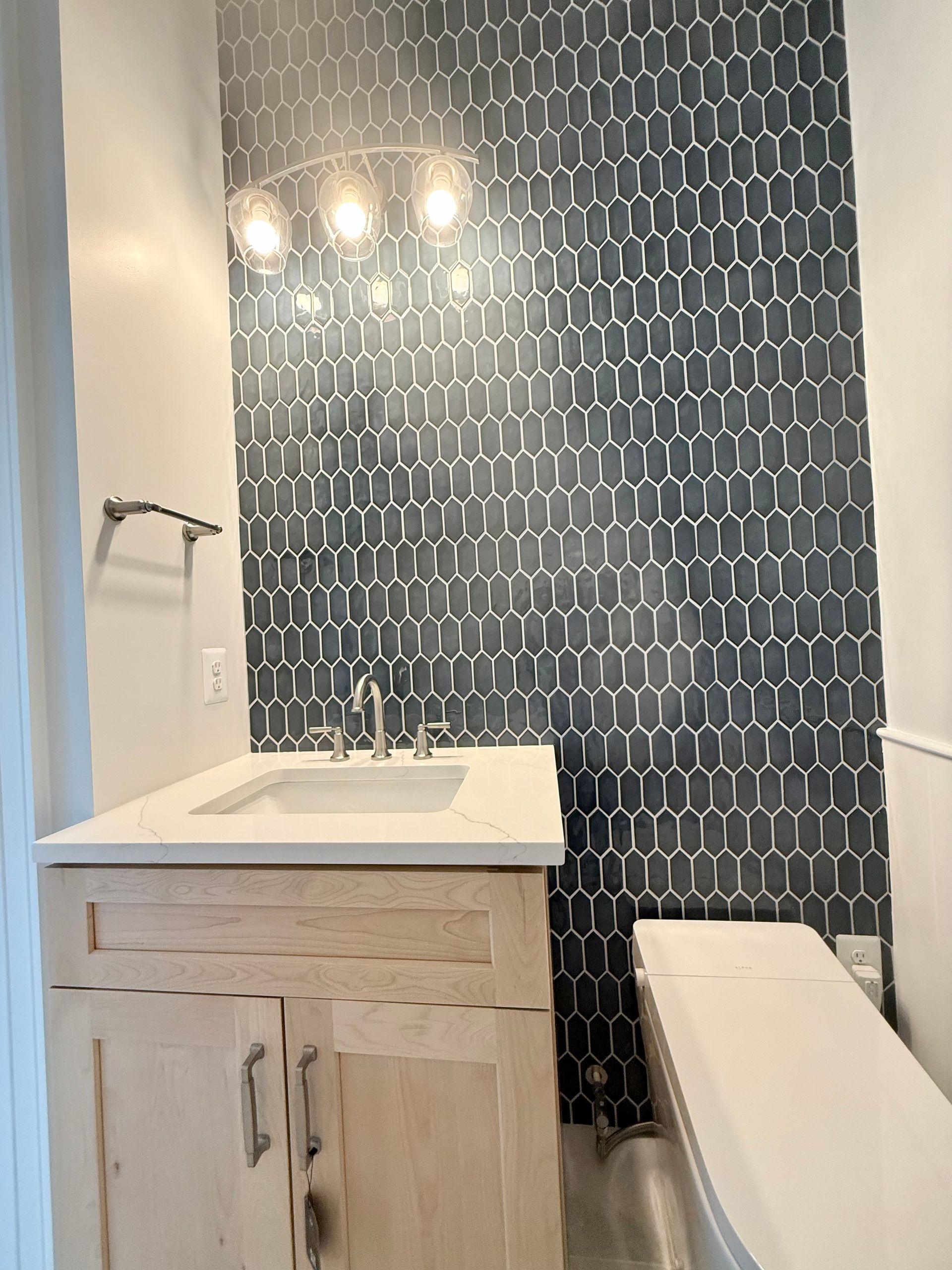 A full view of a powder room remodel showing the bold hexagonal tile accent wall, a natural wood vanity, and a new smart toilet.