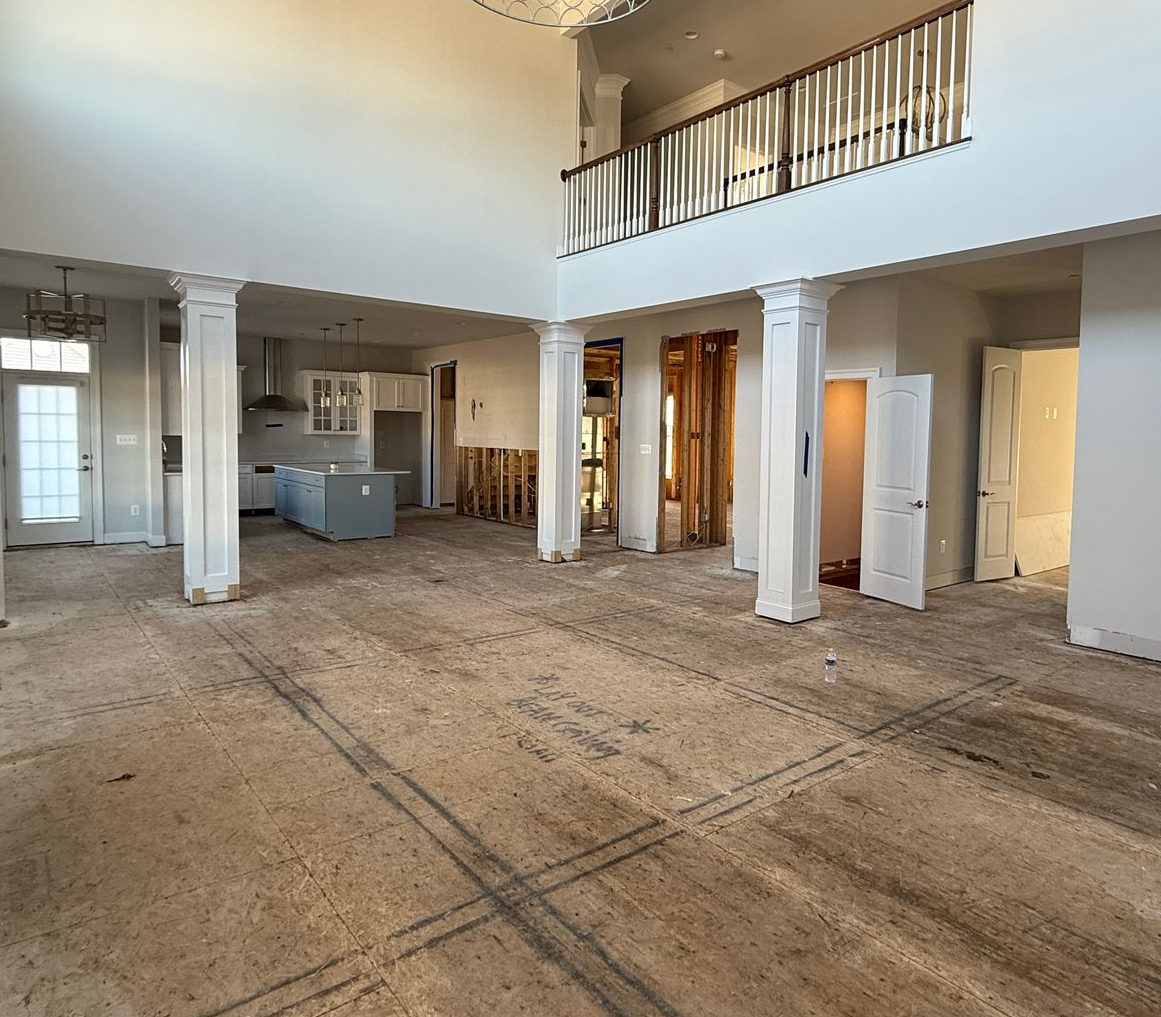 An unfinished living room interior during a remodel, showing exposed walls and subflooring before new wainscoting and hardwood floors.