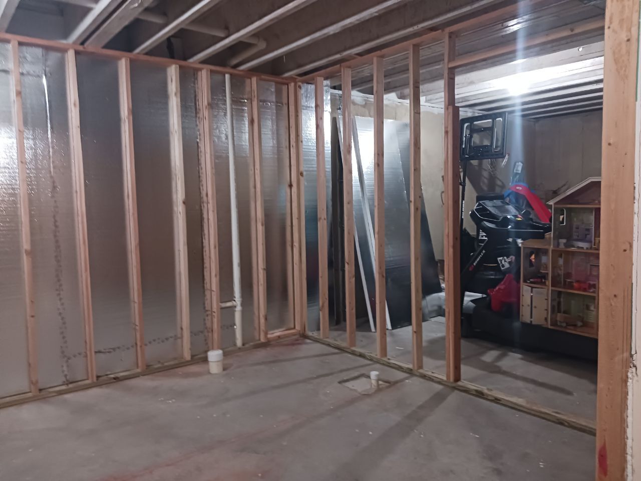 Unfinished basement with exposed wood framing, insulation, and concrete floor, showing the initial state before the bathroom renovation.