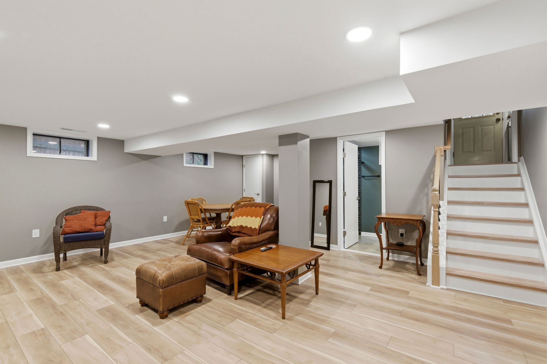 A bright, completed basement living area with light wood-look flooring, smooth gray drywall, recessed lighting, and a large wall-mounted TV.