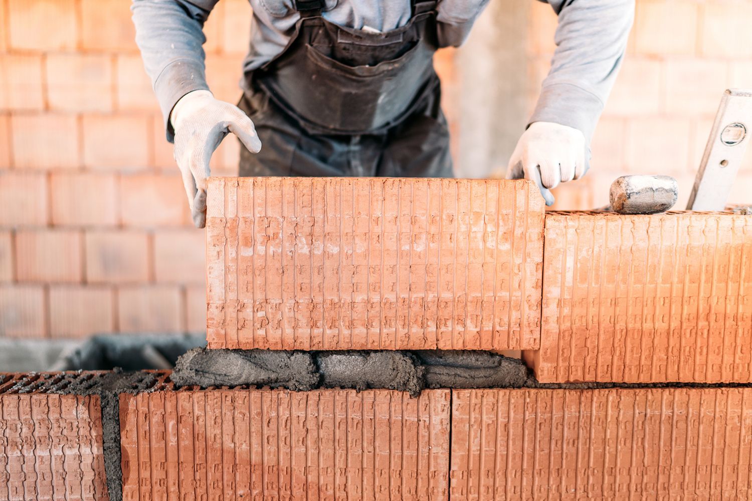 Mason laying a brick on a partially built wall, wearing overalls and gloves.