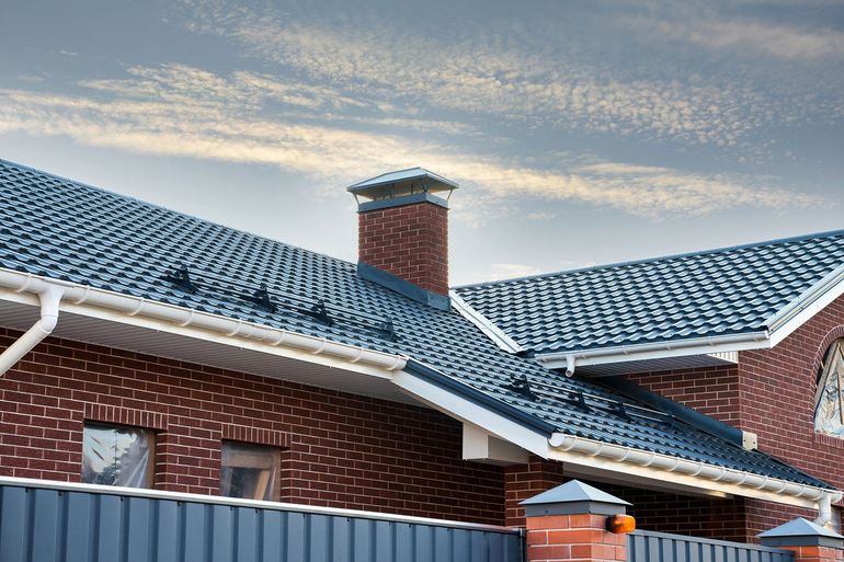 Brick house with blue tiled roof, chimney, white gutters, and blue fence against a cloudy sky.
