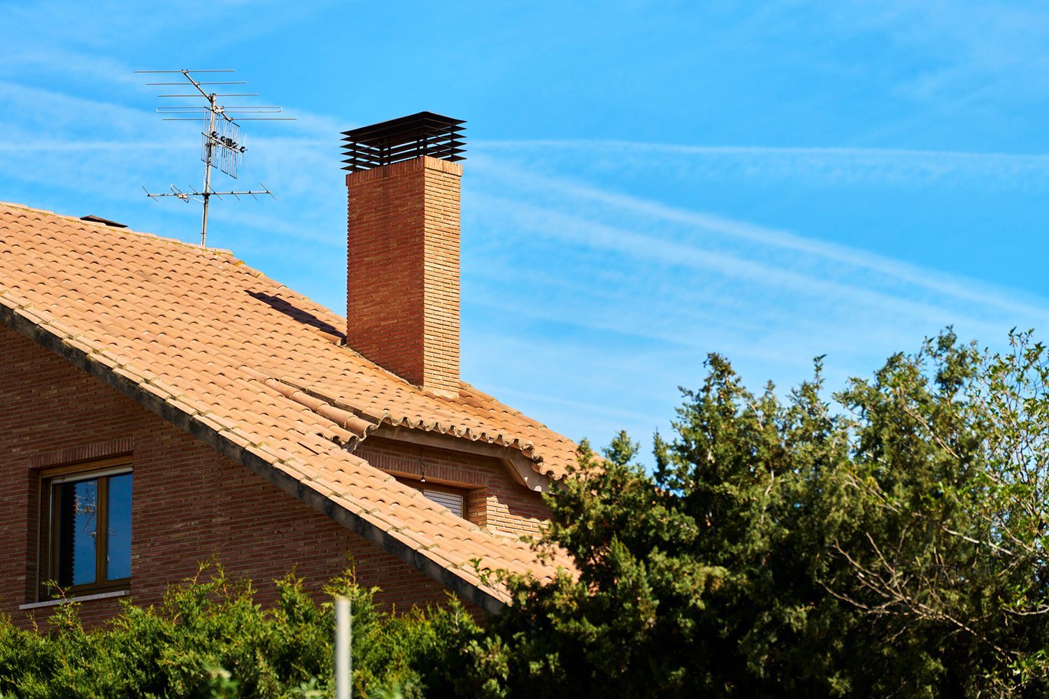 Brick house roof with chimney, antenna, and green trees against a blue sky with cloud trails.