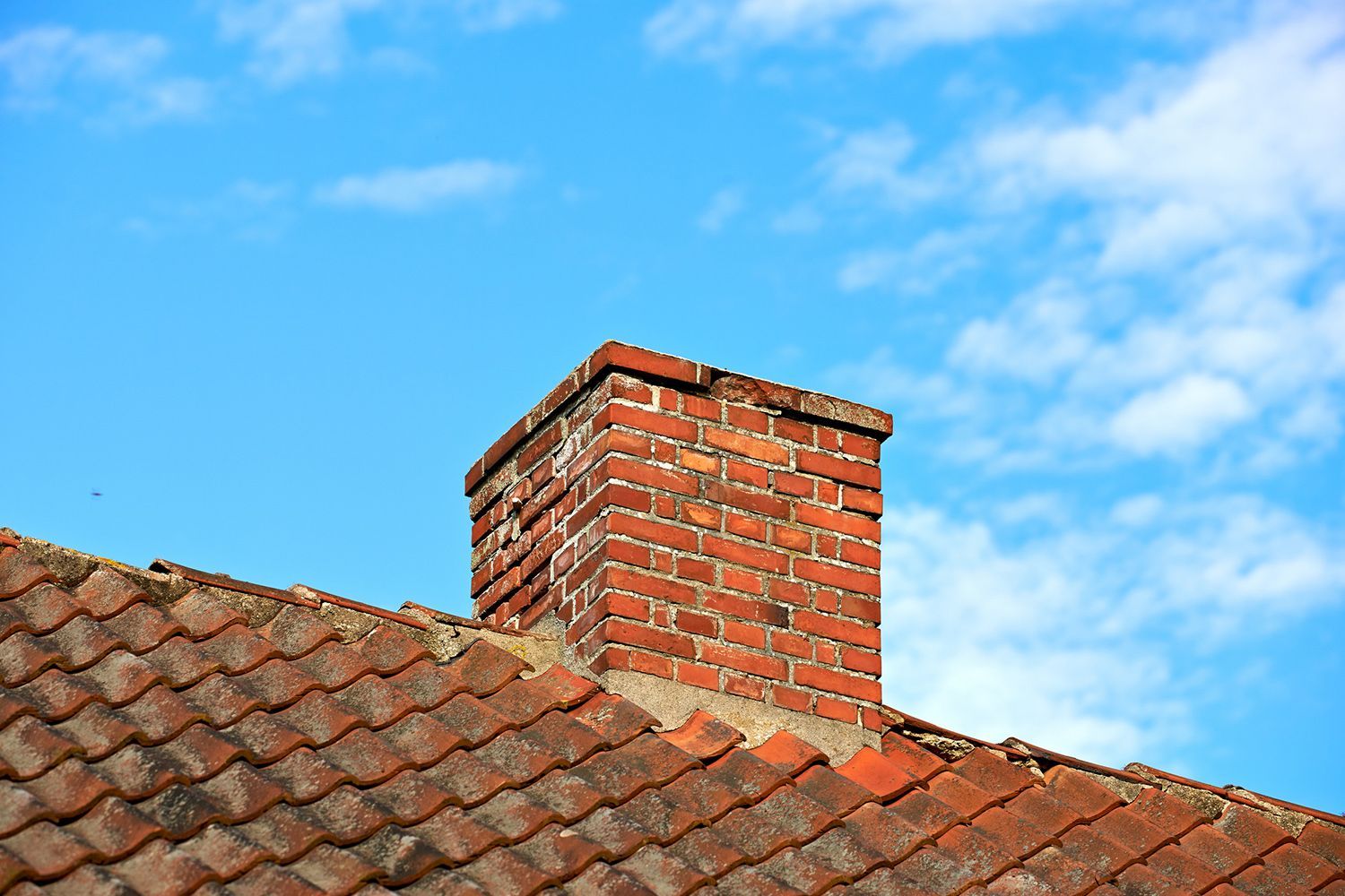 Red brick chimney on a red tiled roof against a bright blue sky with wispy clouds.