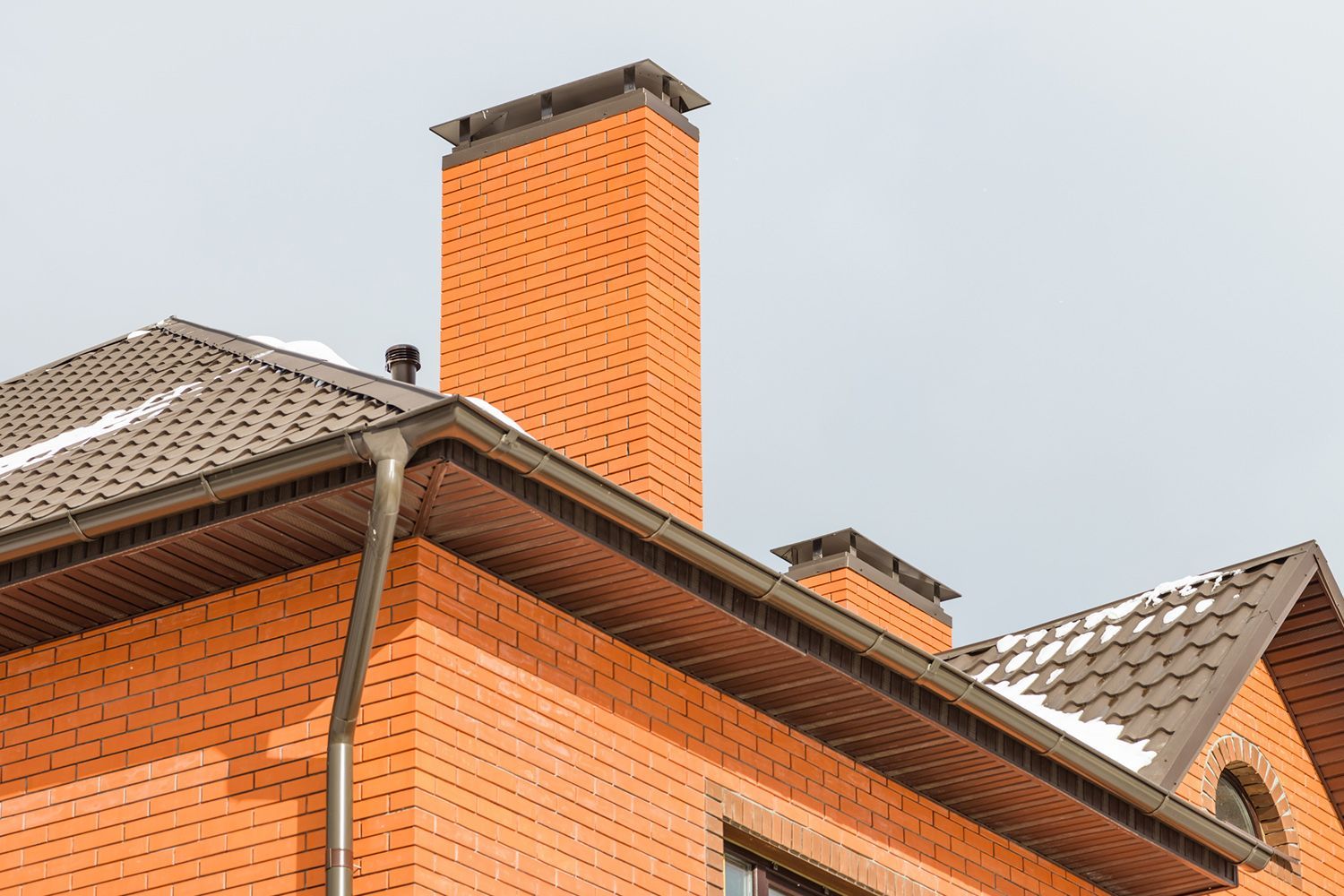 Brick house with brown roof, gutters, and chimney against a cloudy sky.