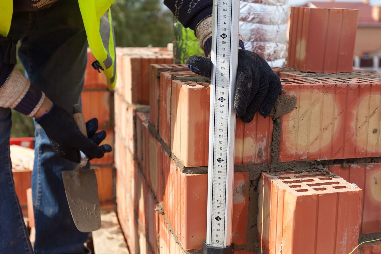 A mason using a level and trowel to build a red brick wall.