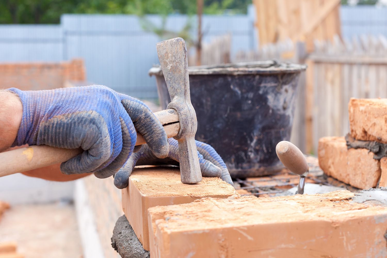 A gloved hand laying bricks with a hammer and mortar in an outdoor construction setting.