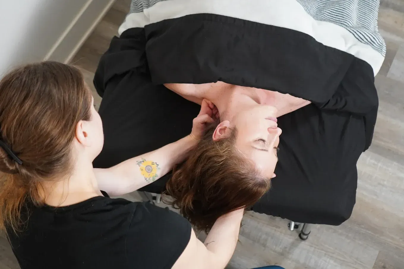 Woman receiving massage on neck; masseuse with arm tattoo. Black drape on massage table.