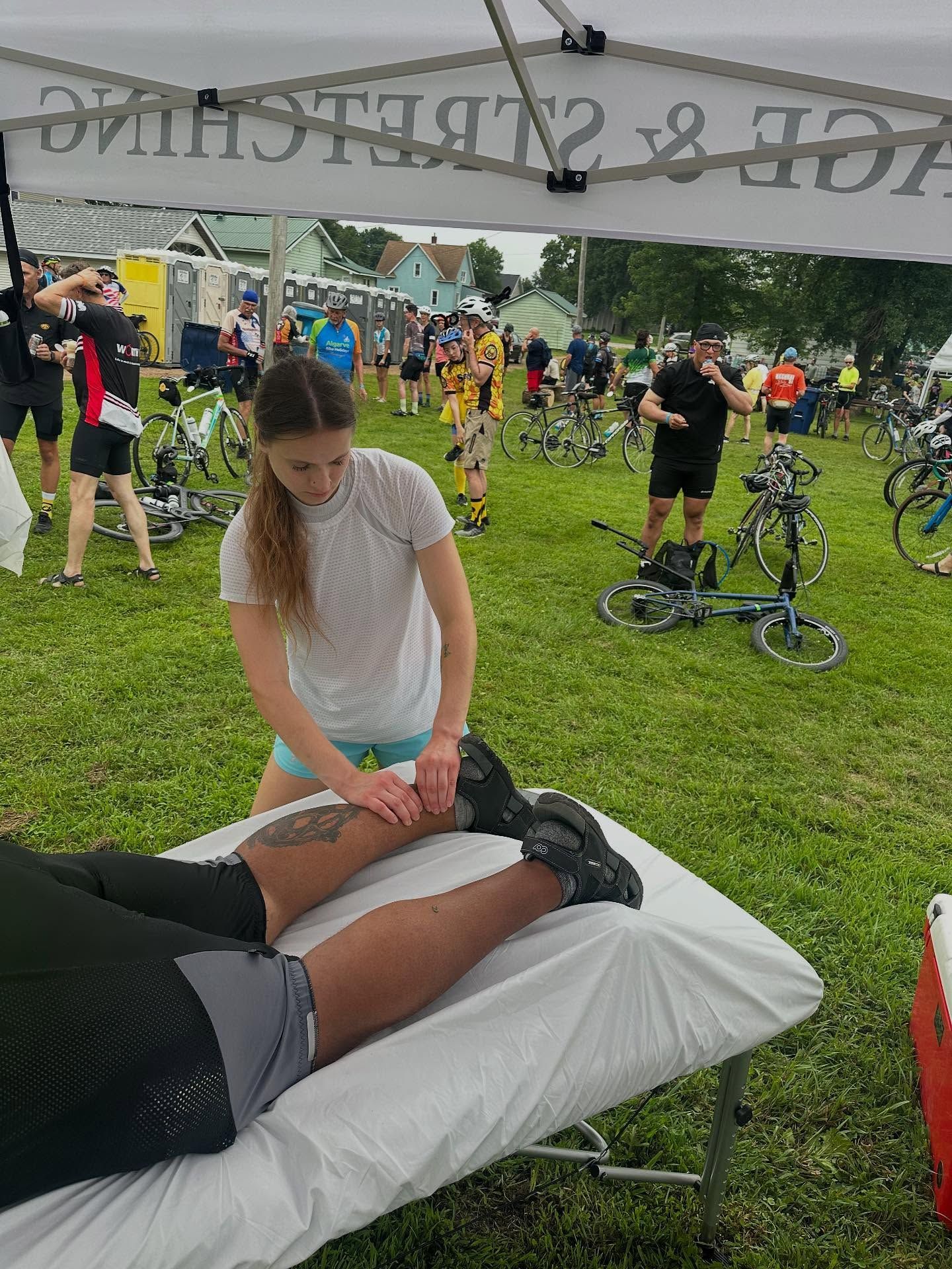 Woman giving leg massage to person on a massage table outdoors, under a tent that reads