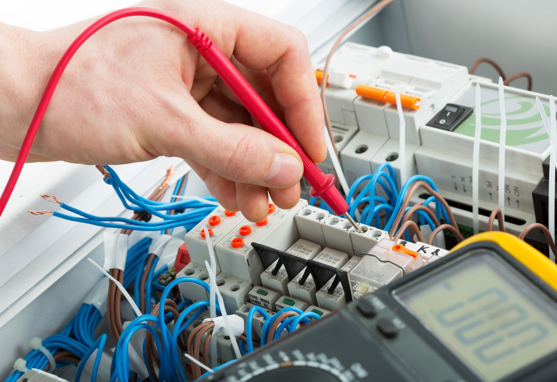 Hand connecting red probe to an electrical test board with wires and a multimeter display